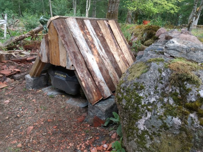 Ett hemmabyggt robotgarage med rustikt träskalstak stödd på stenar i en skogsmiljö.