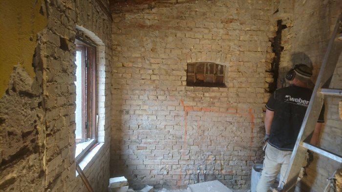 Renoveringsprojekt av ett tegelhus, visar en man som arbetar i ett rum med synliga tegelväggar och partiell putsskada.