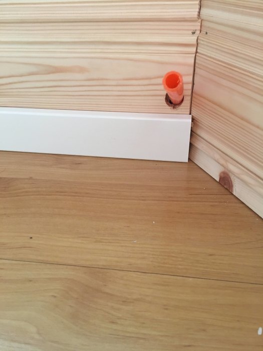 Ett orange tomt rör sticker ut från ett hörn där en vit golvlist möter en ljus trävägg.