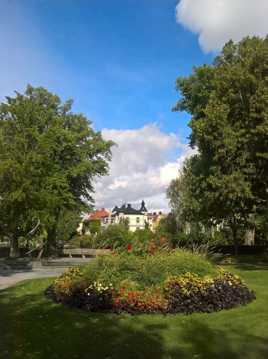 Solig vy av en blomsterprydd park i Wadköping med historiska byggnader i bakgrunden.
