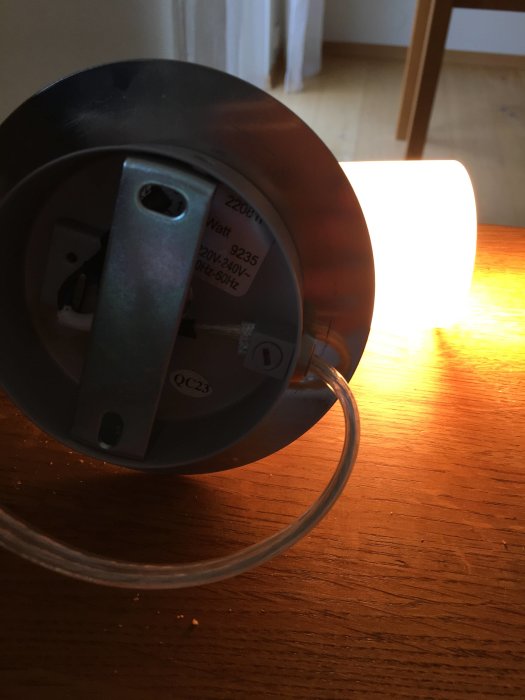 Baksida av lampa med synliga sladdanslutningar och tänd glödlampa i bakgrunden.