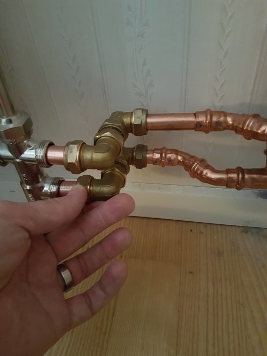 En hand visar nyinstallerade mässingskopplingar och kopparör nära en vägg med vitt, dekorativt tapetmönster.