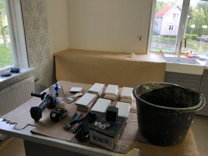 Ett kök under renovering med byggmaterial, verktyg och kakelplattor utspridda på en täckt köksö.