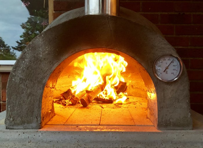 Hembyggd pizzaugn i brand med termometer som visar över 400 grader, sprickor synliga.