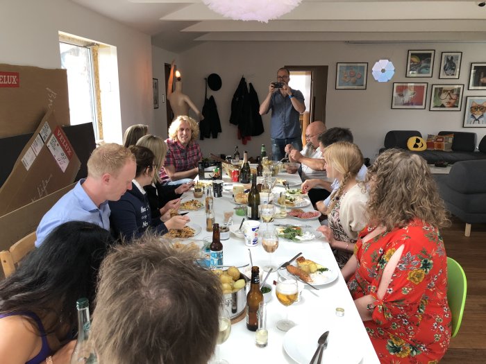 Folk samlade runt matbord inomhus för midsommarfest, mat och drycker på bordet.