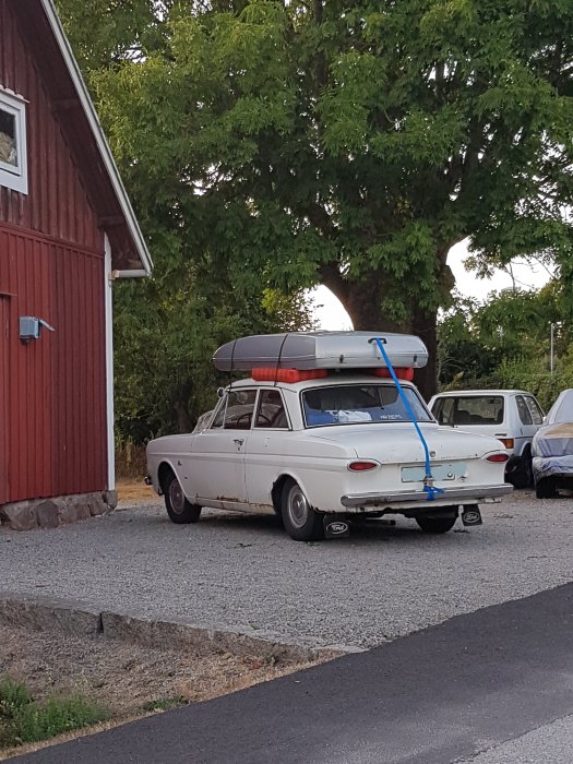 Vit äldre bil med en kanot fastspänd på taket parkerad bredvid en röd stuga.