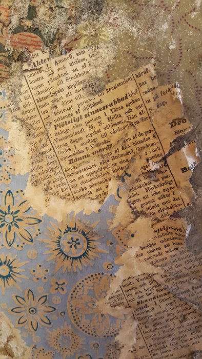 Gammalt tidningspapper delvis täcker en blommig tapet i nyanser av blått och gult, som visar ålderns patina och historik.