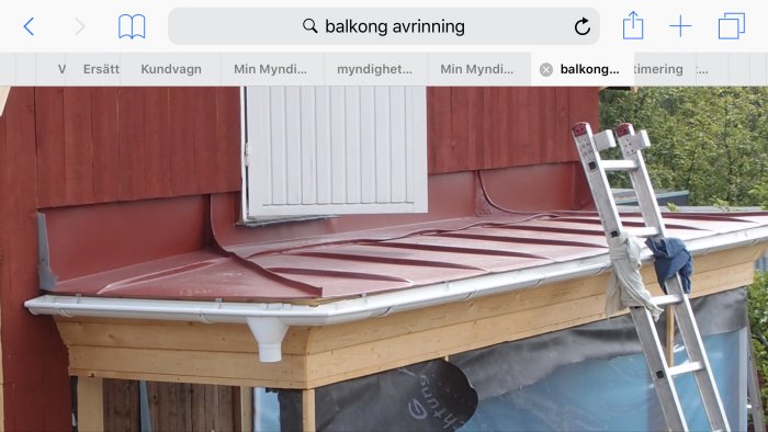 Balkong med röd täckning och vita stuprör under renovering, stege lutar mot sidan.