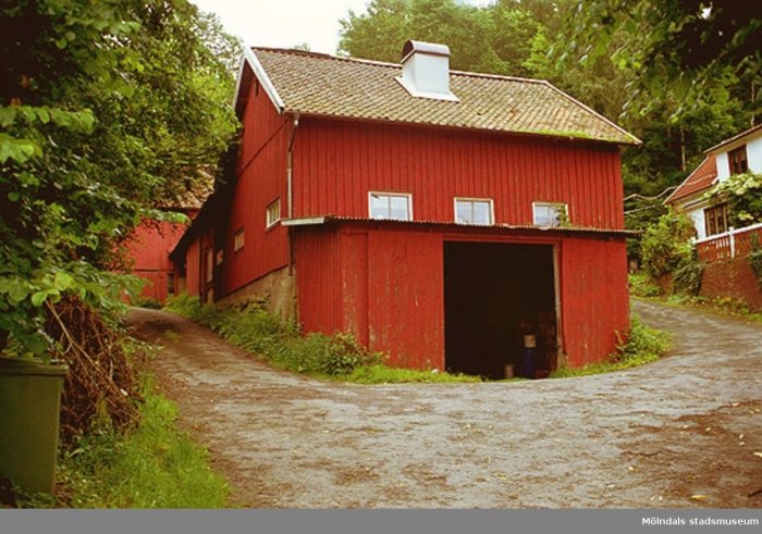 Röd lada med öppen dörr och pulpettak, intill en grusväg och grönska, utan synligt takutsprång.