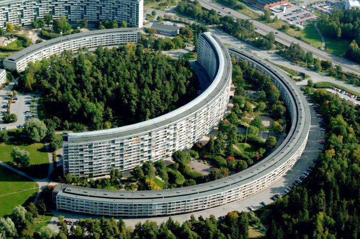 Flygbild över det böjda höghuset i Grindtorp, Täby, omgivet av träd och vägar.