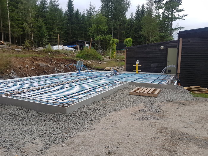 Installation av rör och golvvärme på en ny betongplatta för byggprojekt, omgivet av skog och skjul.