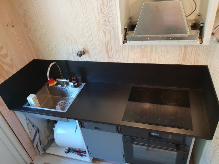 Kök med svart bänkskiva, infälld spishäll, diskho och plywoodvägg, visar skarv mellan paneler.