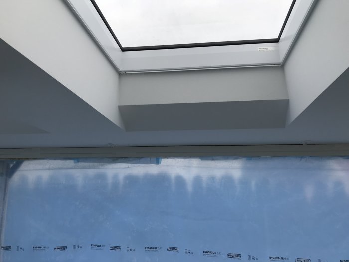 Takfönster med vinklade smygar och en vägg täckt av byggplast med isoleringsmaterial synlig.