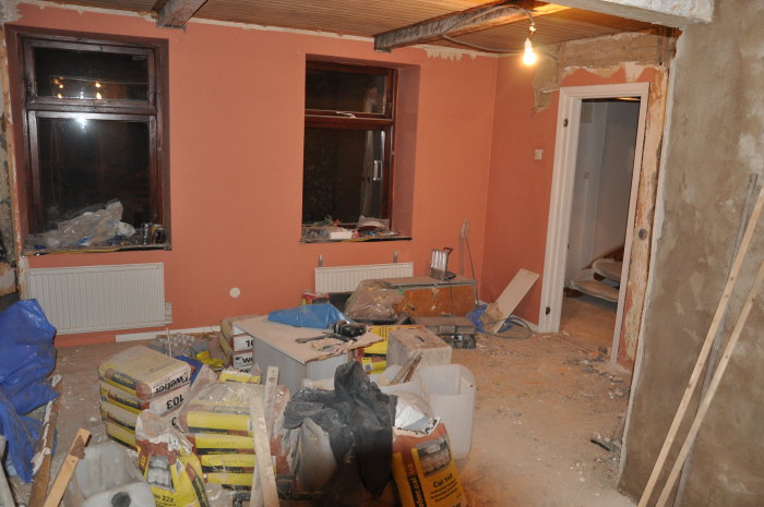 Renoveringsarbete i vardagsrum med exponerad isolering, uppbrutna golv och byggmaterial överallt.