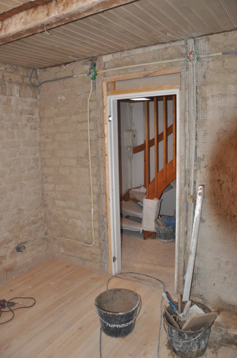 Renoveringsprojekt i vardagsrum med oslipade murväggar, exponerade kablar och nytt trägolv, genomgång till annat rum syns.