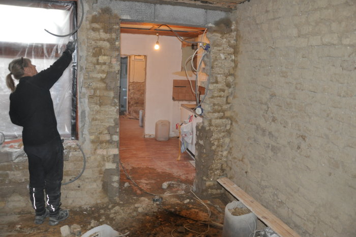 Renoveringsarbete i ett vardagsrum med synliga tegelväggar och ny eldragning, där en person arbetar med väggstrukturen.