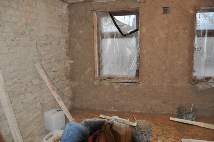 Renoveringsarbete med lerputsad vägg, byggmaterial och verktyg framför två fönster skyddade med plast.