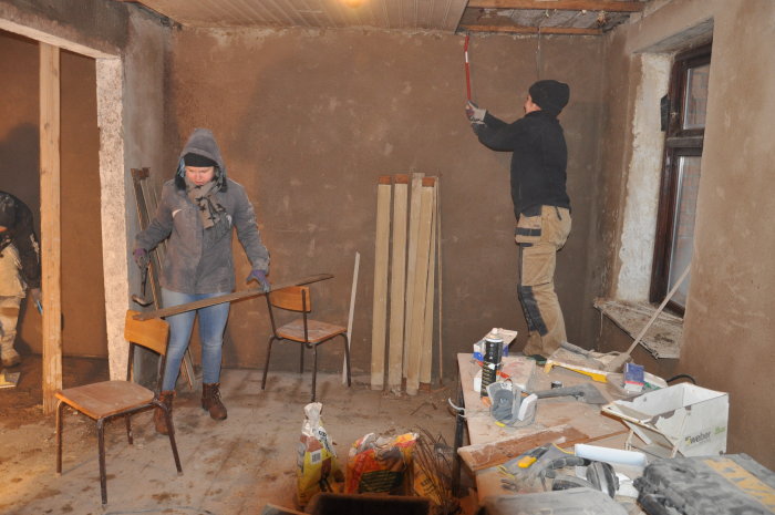 Två personer renoverar ett rum, en målar vägg och den andra bär stolar, med byggmaterial runtomkring.
