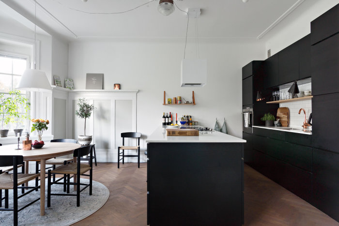 Modernt kök med svarta skåp, vit köksö, inbyggda vitvaror och matplats med träbord och stolar.