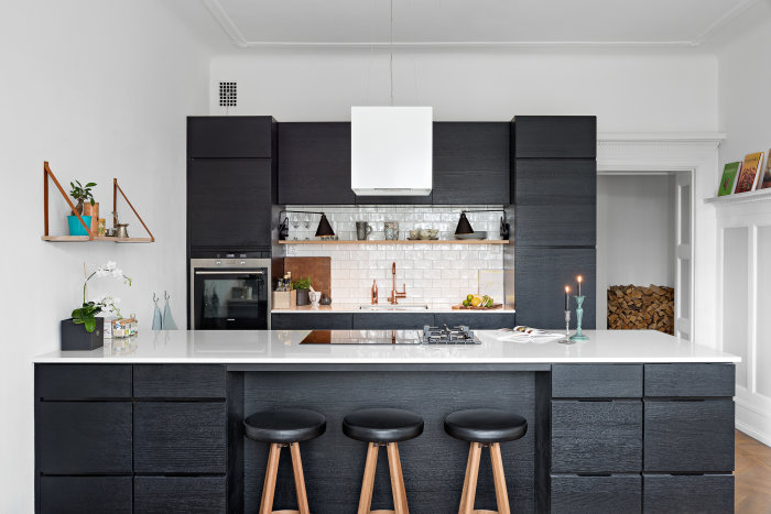 Modernt kök med svarta skåp, vit bänkskiva, köksö, inbyggd ugn, gasspis och hängande lampor.
