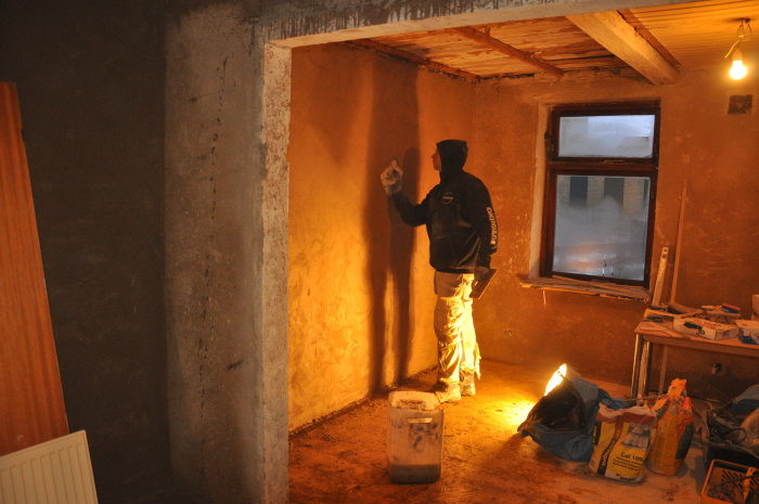 Person applicerar kalkgrund på en vägg i ett renoveringsrum med byggmaterial synliga.