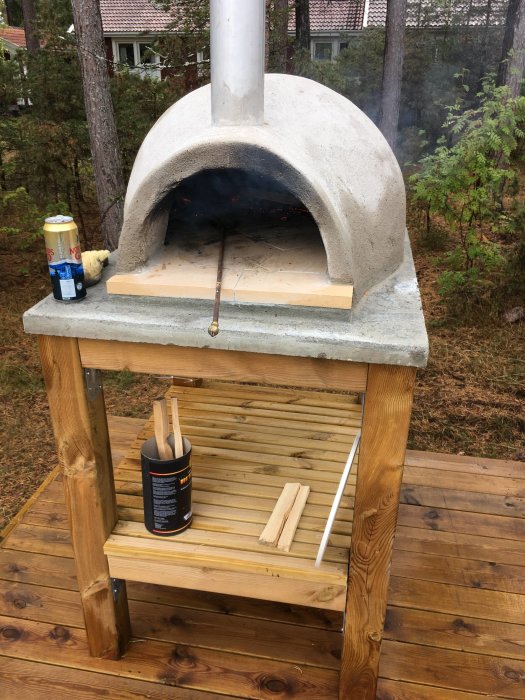 Utomhus vedeldad pizzaugn av betong på trädäck med ved och tillbehör.