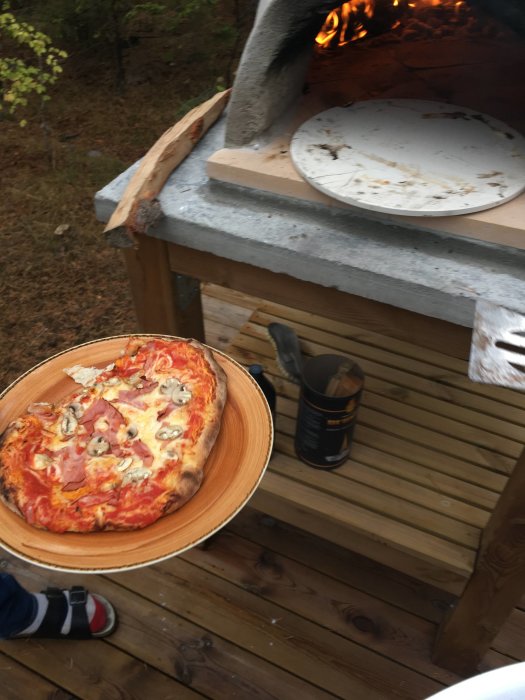 Nybakad pizza på träbricka framför en utomhuspizzaugn med eld och en tom pizzasten inuti.