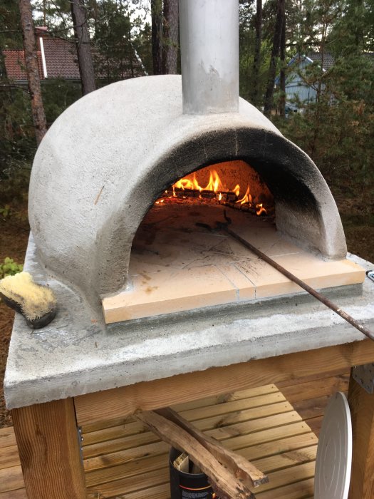 Utomhus pizzaugn av sten med eld i bak och pizzastenar framför öppningen.