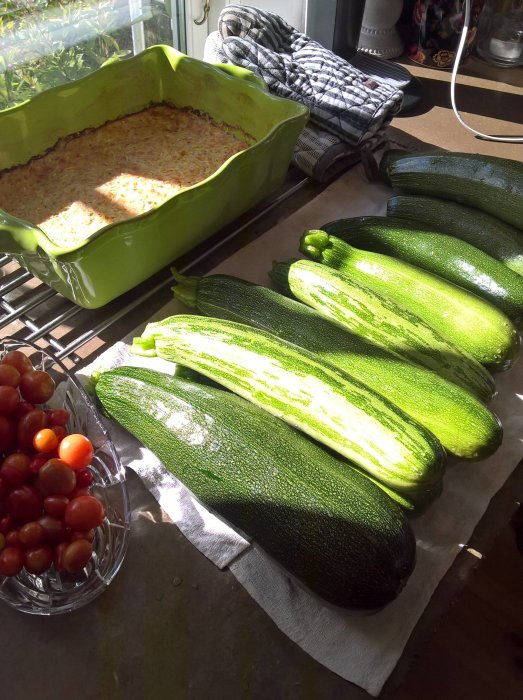 Stora gröna squash och en skål med röda tomater på ett köksbord, bakad maträtt i en grön form i bakgrunden.