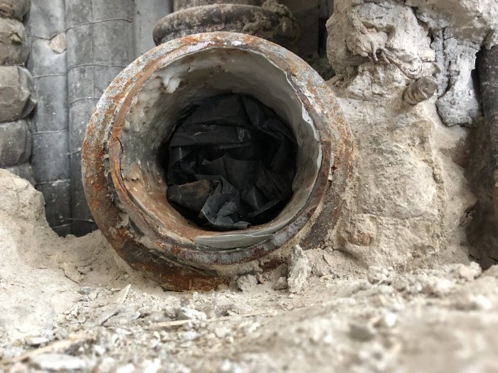 Öppet schakt med synligt gjutjärnsavloppsrör täckt med svart plast, omgivet av betong och rörledningar.