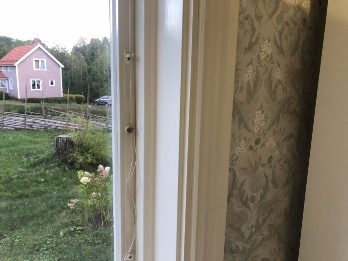 Fönster med nysatt vitt foder i ett hem, med utsikt över en trädgård och ett rosa hus.