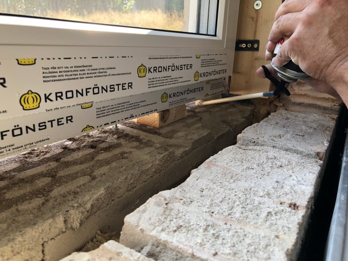 Installation av nytt fönster med fogskum, synlig Kronfönster-logotyp och en hand som applicerar skummet längs fönsterkarmen.