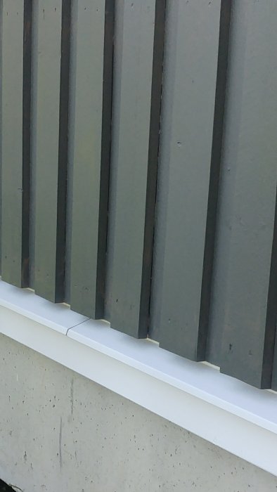 Vägg med lodräta paneler ovanför vit vattenavrinning utan droppnäsa, vid grå betonggrund.