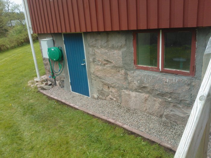 Husgrund med lager av 8-16 grus längs väggen, röd takplåt, blå dörr och en grön vattenslang.
