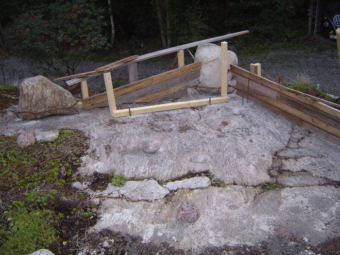 Förberedelser för altanbygge med hörnstenar och armeringsjärn på en bergig grund.