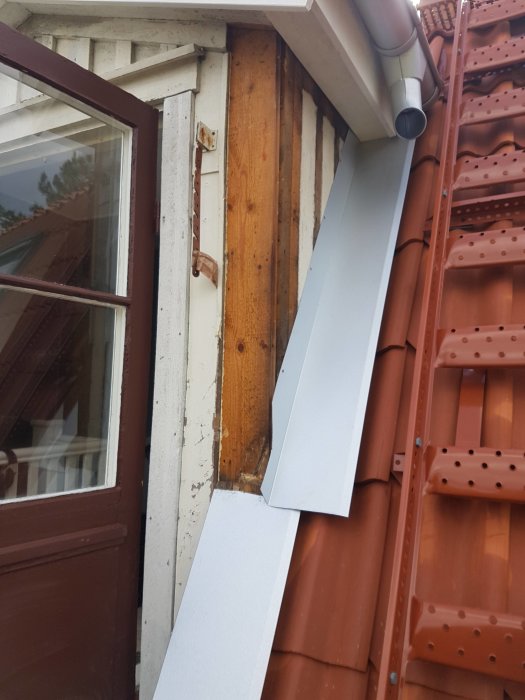 Bild på en hörna av ett hus med röda takpannor, vita plåtar och synlig träpanel vid takkupan.