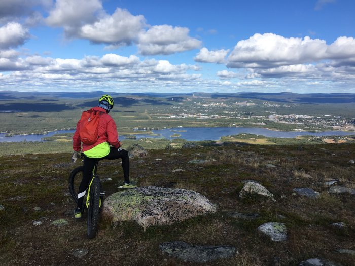 Cyklist på en elcykel som betraktar utsikten över ett landskap med sjöar och en stad i fjärran från ett berg.