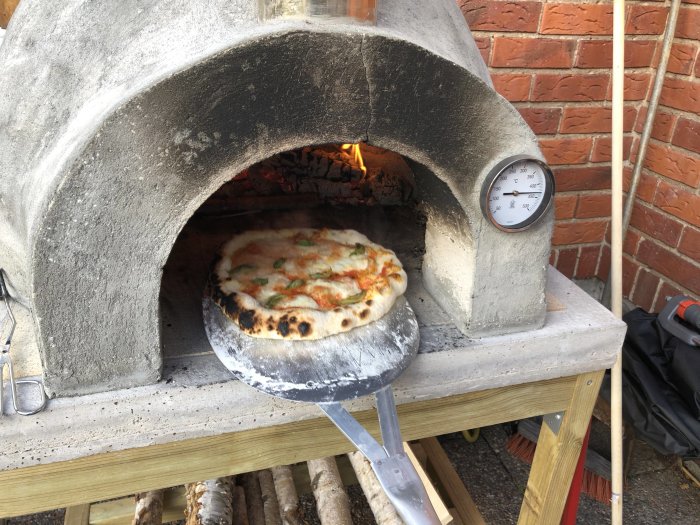 Pizza tas ut ur en vedeldad ugn med en temperaturmätare som visar 450 grader.