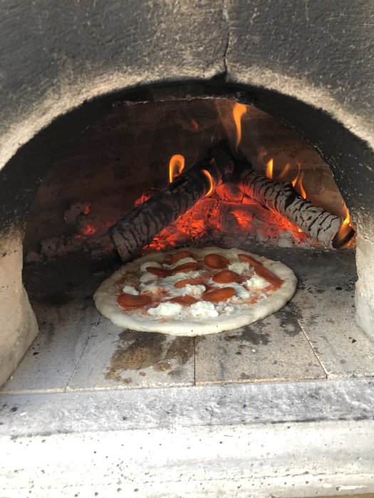 Pizza gräddas i en vedeldad ugn med synliga lågor och glödande ved.