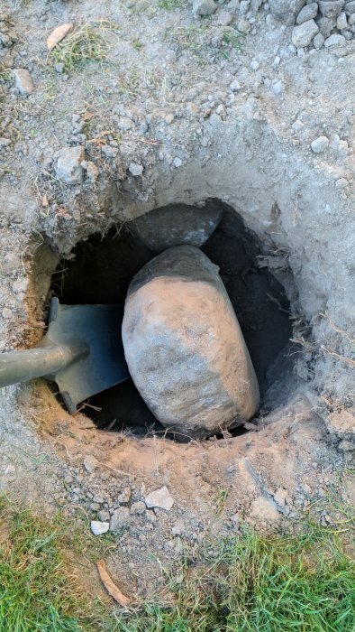 Spade som är i kontakt med en stor sten i en grävd grop under plintgrävningsarbetet för altanbygge.