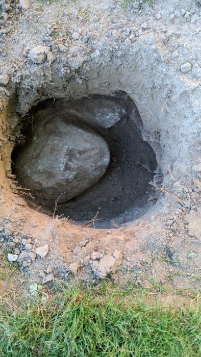 Ett grävt hål i marken visar en stor sten som förhindrar nedkörning av markskruv, omringad av jord och småsten.
