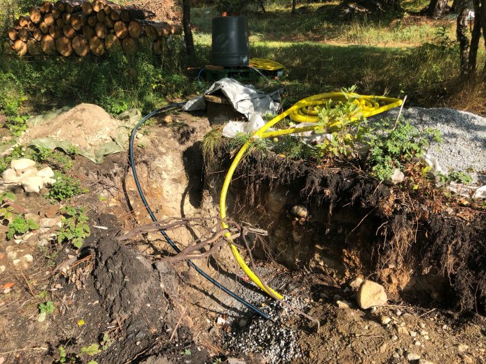 Grävd brunn med synliga PEM-slangar och kablar omgivna av grus och vegetation under en renoveringsprocess.