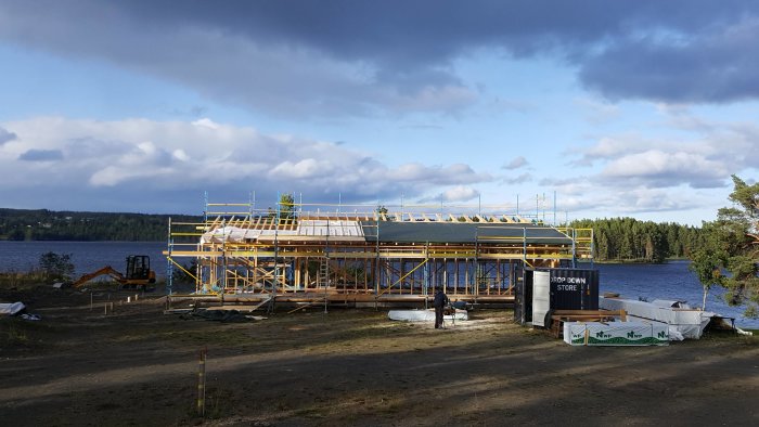 Enplansvilla under konstruktion på udde vid sjö med byggmaterial runtomkring och ställningar runt byggnaden.