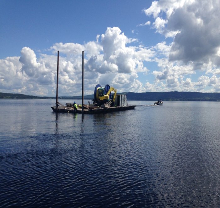 Arbetsflotte med utrustning för läggning av sjökabel på en klarblå sjö med fluffiga moln i bakgrunden.