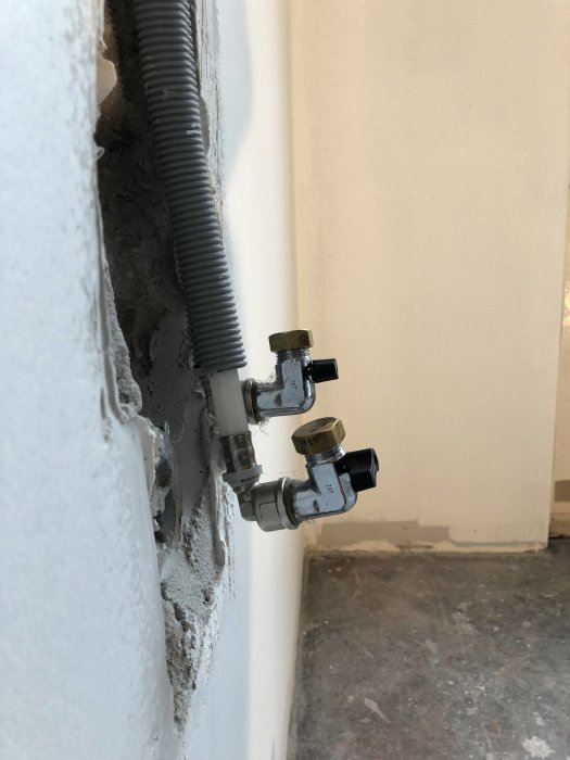 Nyinstallerade vattenrör och ventiler som sticker ut ur en vitmålad vägg och ett oslipat betonggolv.
