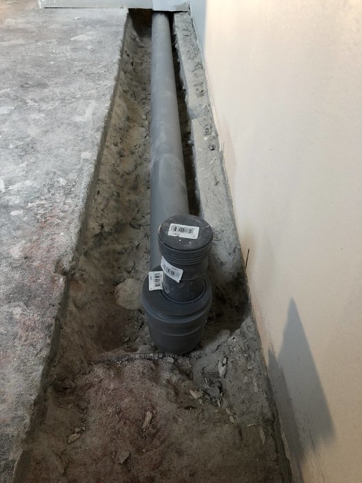 Grått avloppsrör i grävd ränna längs en betongvägg i ett renoveringsprojekt.