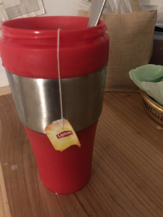 Röd termosmugg med Lipton tepåse hängande över kanten och en sked i.