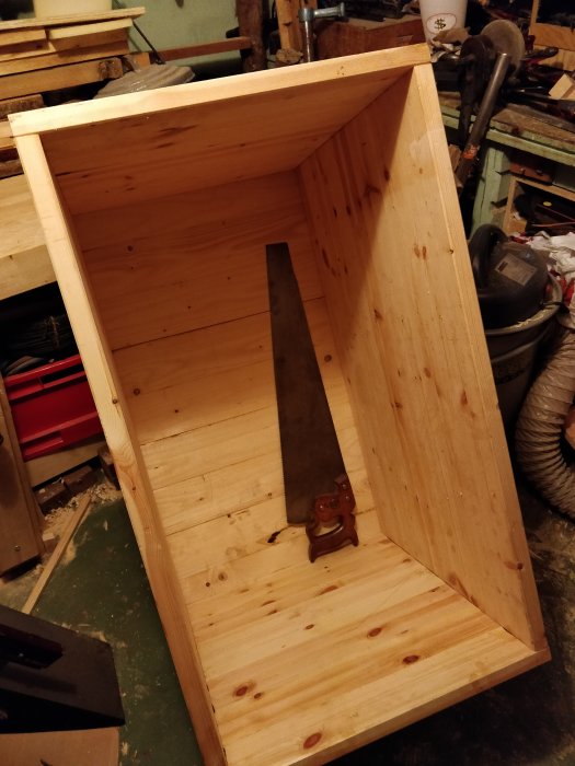 Hemmabyggd trälåda med nygjort golv och en stor handsåg inuti, i en verkstadsliknande miljö.