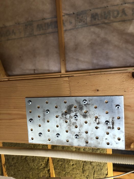 Metallplatta fastsatt på träbjälke med murarsnöre framför, i en byggkonstruktion med isoleringsmaterial i bakgrunden.