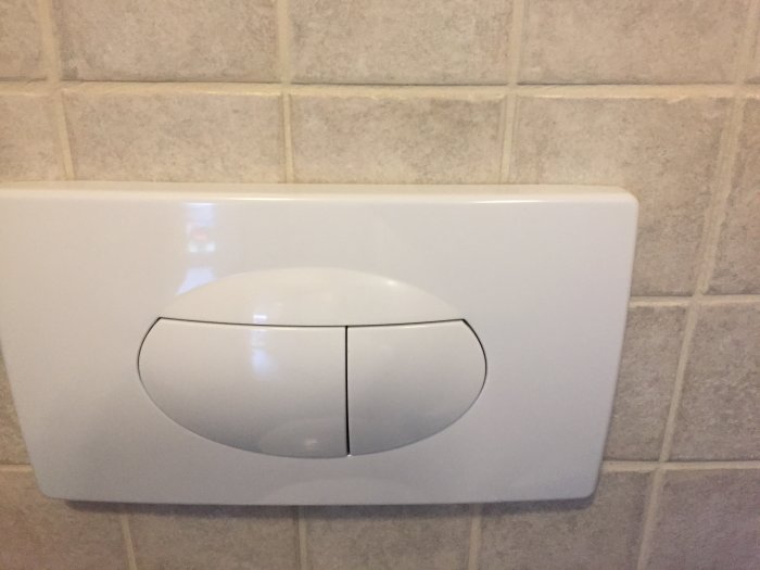 Vit vägghängd WC-spolknapp med två knappar monterad på kakelvägg.